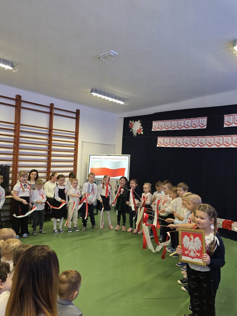 Grupa dzieci w białych koszulach z czerwonymi kokardami stoi na tle ciemnej ściany z polskimi flagami. Dzieci trzymają biało-czerwone wstążki, jedna z nich trzyma herb Polski. Tło jest ozdobione plakatami o Święcie