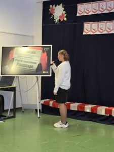 Osoba prezentuje slajd o Piłsudskim na tle ciemnej kurtyny z różami. Na ekranie duży tekst gazety. Podłoga jest zielona.