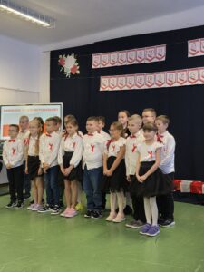 Grupa dzieci w strojach galowych stoi na zielonej podłodze. Czarne tło z napisem Narodowe Święto Niepodległości. Dekoracja z kwiatów po lewej stronie.