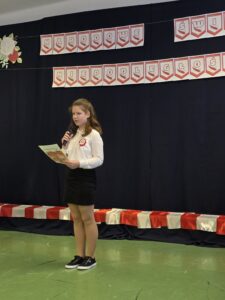 Dziewczyna w białej bluzce przemawia na tle ciemnej kurtyny z napisem Narodowe Święto Niepodległości. Dekoracje biało-czerwone.