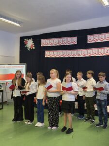 Grupa dzieci w białych koszulach z książkami stoi na tle czarnego plakatu. Na nim jest napis Narodowe Święto Niepodległości. Podłoga jest zielona.