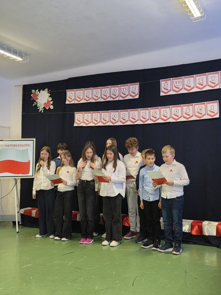 Grupa dzieci w białych koszulach śpiewa przed czarnym tłem z napisem Narodowe Święto Niepodległości. Widoczne biało-czerwone dekoracje i plansza z flagą Polski. Podłoga jest zielona.