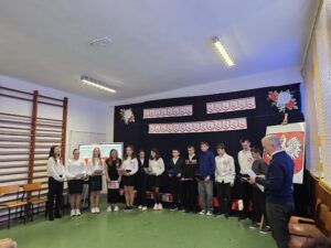 Grupa uczniów i dorosłych stoi na zielonej podłodze szkolnej sali. Tło zdobią biało-czerwone dekoracje. Prawa strona zawiera herb Polski na sztaludze.
