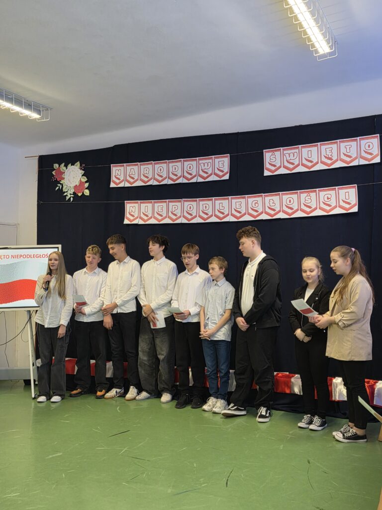 Grupa uczniów stoi na tle czarnego koca ozdobionego napisem Narodowe Święto Niepodległości. Ubrani w białe koszule, trzymają kartki w rękach. Na podłodze jasne, zielone pokrycie.