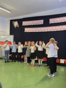 Dzieci w białych koszulach i spódniczkach, występują na scenie. Czarne tło z napisem Narodowe Święto Niepodległości. Podłoga w kolorze zielonym.