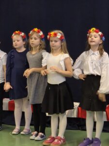 Cztery dziewczynki w wiankach z kwiatami stoją na scenie. Ubrane w sukienki i bluzki, tło jest ciemne. Obok znajduje się biało-czerwona dekoracja.