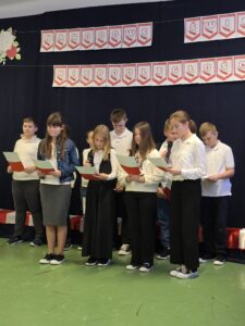 Grupa dzieci w białych koszulach stoi na zielonej podłodze. Trzymają kartki i mikrofony, na tle czarnej kurtyny z napisem Narodowe Święto Niepodległości.