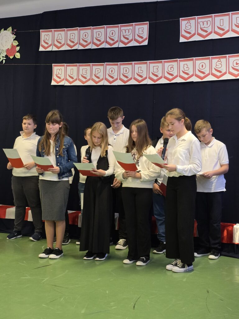 Grupa dzieci w białych koszulach stoi na zielonej podłodze. Trzymają kartki i mikrofony, na tle czarnej kurtyny z napisem Narodowe Święto Niepodległości.