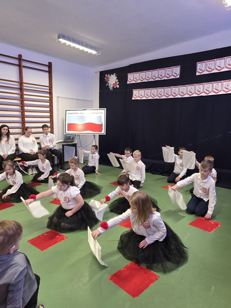 Dzieci w białych bluzkach i czarnych spódniczkach trzymają białe chustki, siedzą na ziemi. Czerwono-biały wystrój ściany. Tło zielone z czerwonymi akcentami.