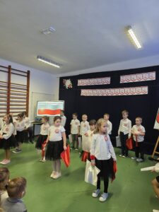 Grupa dzieci w białych koszulkach i czarnych spódnicach na tle dekoracji z napisem Narodowe Święto Niepodległości. Dzieci trzymają czerwono-białe flagi. Sala ma zieloną podłogę i drewnianą drabinkę