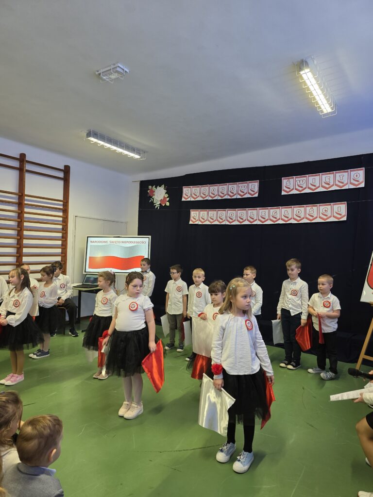 Grupa dzieci w białych koszulkach i czarnych spódnicach na tle dekoracji z napisem Narodowe Święto Niepodległości. Dzieci trzymają czerwono-białe flagi. Sala ma zieloną podłogę i drewnianą drabinkę