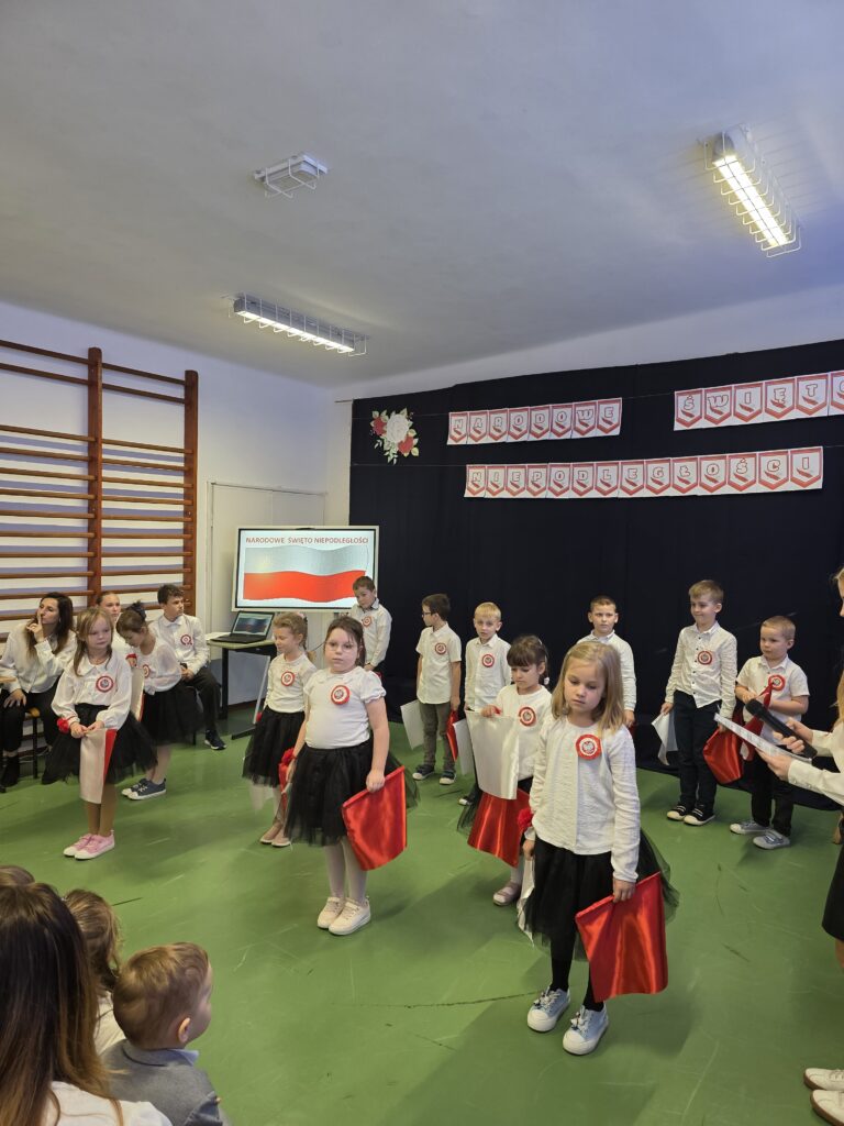 Dzieci w białych bluzkach i czarnych spódniczkach z czerwonymi emblematami stoją na zielonej podłodze. W tle ciemna kurtyna z napisem Narodowe Święto Niepodległości. Po lewo drabinki i ekran z