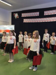 Grupa dzieci w białych bluzkach i czarnych spódnicach trzyma czerwone flagi. W tle czarny transparent z napisem Narodowe Święto Niepodległości. Zielona podłoga stanowi kontrast dla jasnych strojów dzieci.