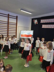 Dzieci w białych koszulach i czarnych spódniczkach trzymają polskie flagi. Tło to ściana z drabiną i symbolem Święta Niepodległości. Dominują kolory biały, czerwony i czarny.