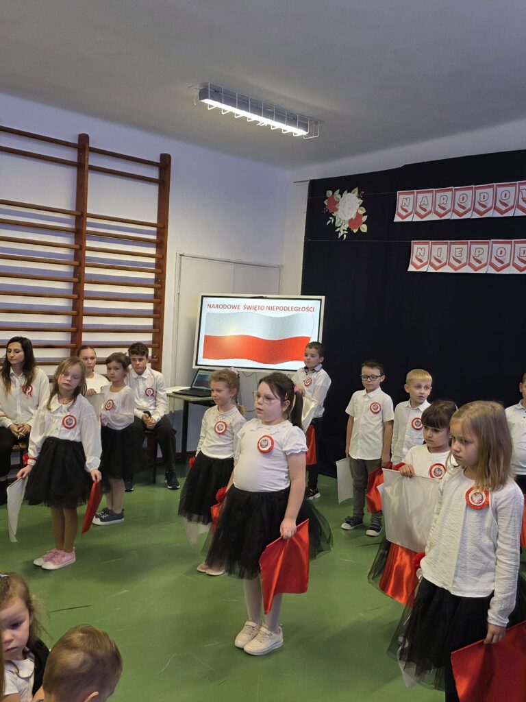 Dzieci w białych koszulach i czarnych spódniczkach trzymają polskie flagi. Tło to ściana z drabiną i symbolem Święta Niepodległości. Dominują kolory biały, czerwony i czarny.