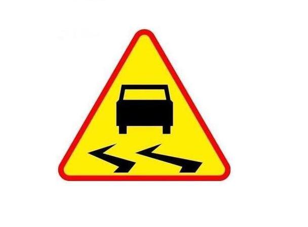 Żółty znak ostrzegawczy z czarnym samochodem i liniami, sugeruje śliską nawierzchnię. Ma czerwoną obwódkę. Tło jest białe.