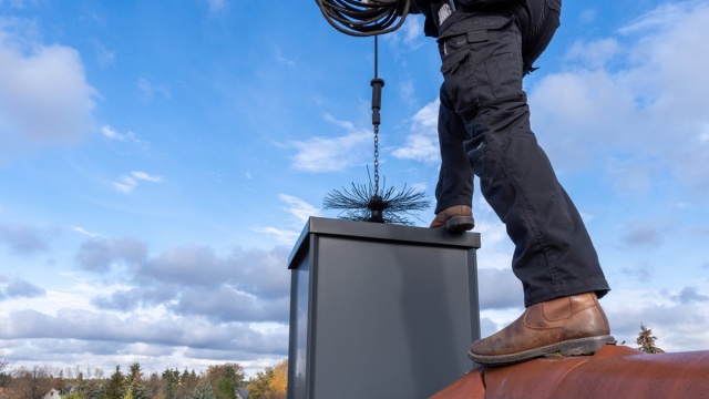 Osoba na dachu czyszczy komin szczotką. Niebo jasnoniebieskie z chmurami. Tło: linia drzew.