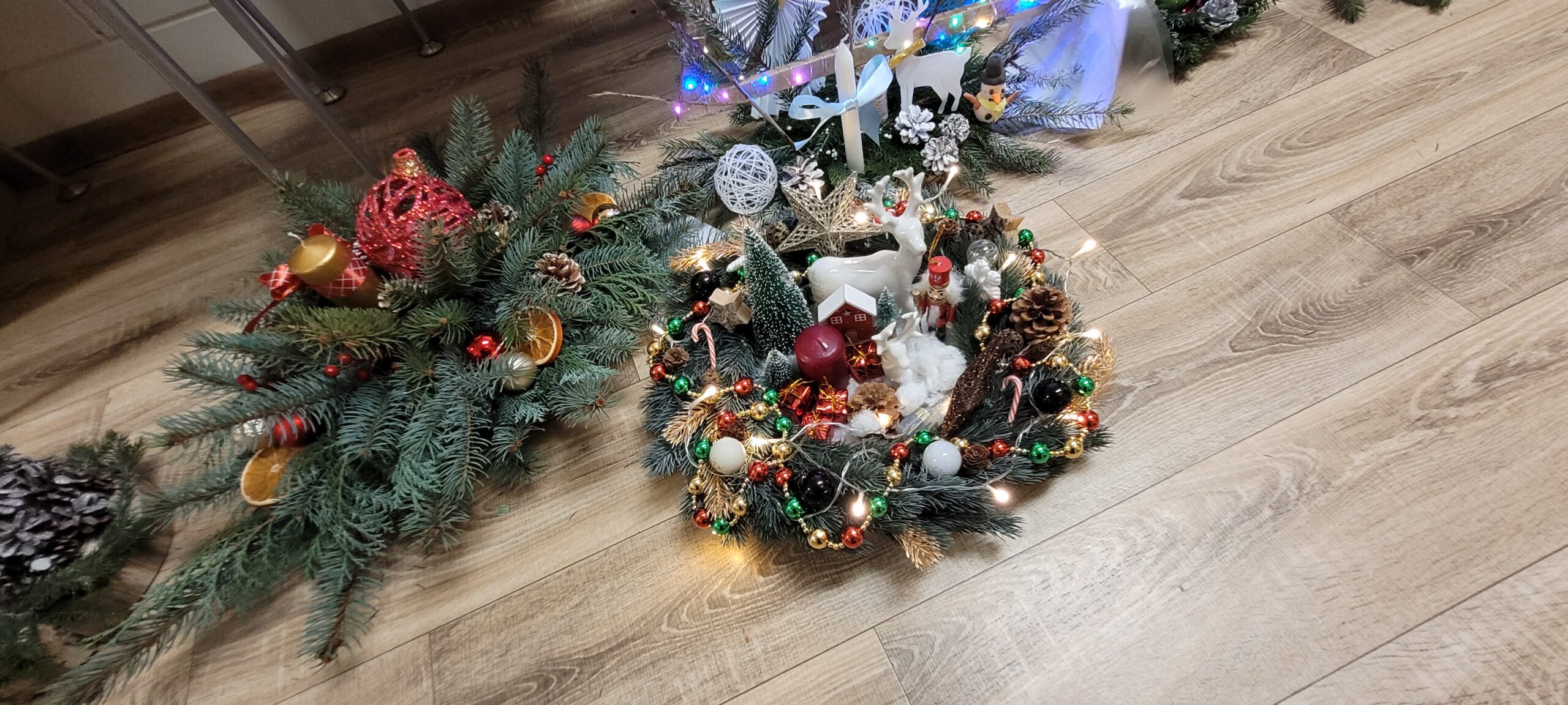 Dwa świąteczne stroiki z gałązkami iglastymi leżą na drewnianej podłodze. Jeden stroik zdobią czerwone bombki i suszone pomarańcze. Drugi stroik ozdobiony jest białymi figurkami jeleni i mniejsze kolorowe bombki.