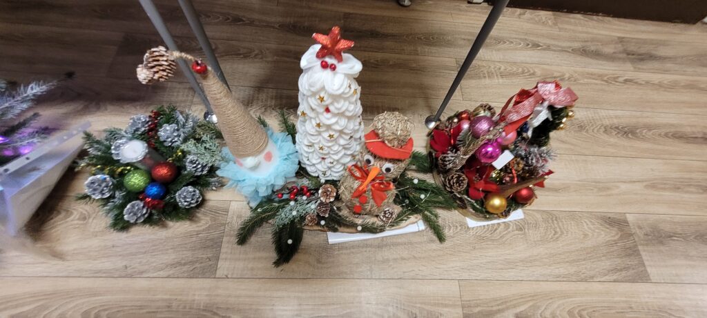 Trzy świąteczne dekoracje na drewnianej podłodze. Jedna to choinka ozdobiona gwiazdą, kolejna to skrzat, a trzecia to bałwan. Kolory to głównie biały, czerwony i zielony z dodatkami błyszczących bombek.