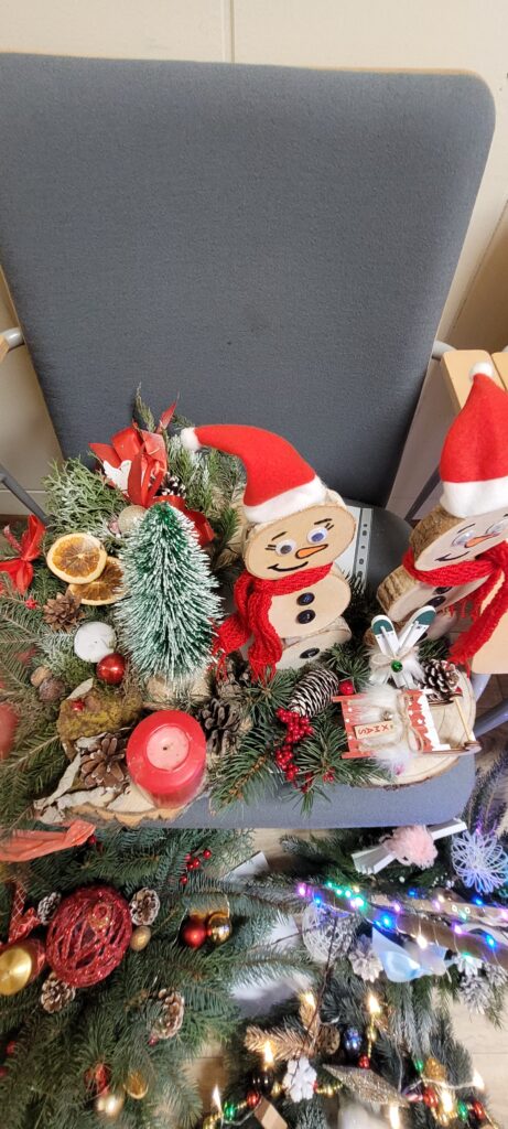 Świąteczna dekoracja na stole z drewnianymi bałwankami i zielonymi gałązkami. Tło szare, stonowane. Dominuje czerwień i zieleń.