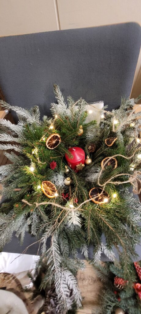 Wieniec z gałązek świerku z czerwoną świecą i złotymi bombkami. Ozdobiony suszonymi pomarańczami i światełkami. Tło szare krzesło.