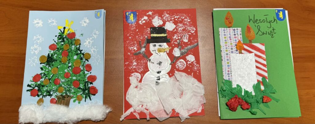 Na zdjęciu są trzy kolorowe kartki świąteczne na biurku. Pierwsza ma choinkę na niebieskim tle, druga bałwana na czerwonym tle, a trzecia świece na zielonym tle z napisem Wesołych Świąt. Kolorowe ornamenty dodają ś