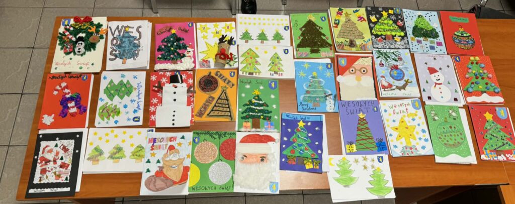 Szereg kolorowych kartek świątecznych leży na drewnianym stole. Na kartkach widoczne są choinki, bałwanki i napisy Wesołych Świąt. Tło stanowi szary kaflowy podłoga.