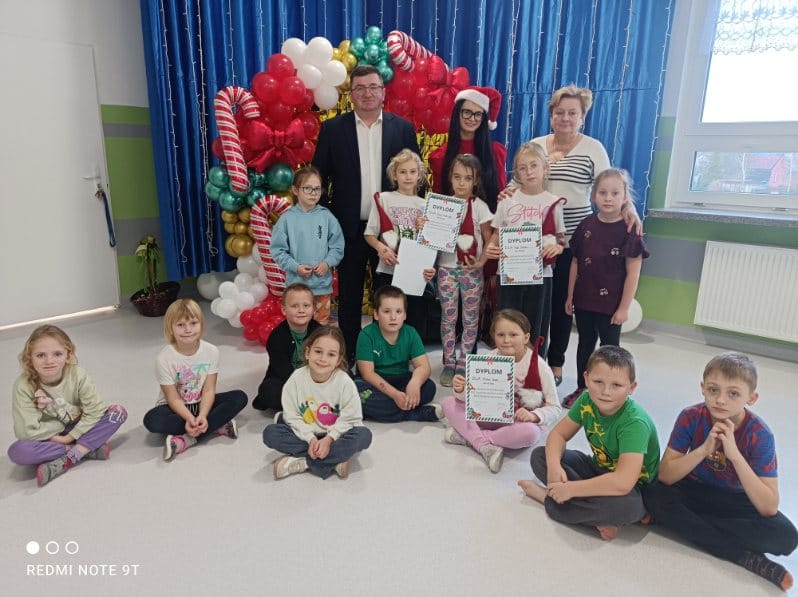 Na zdjęciu grupa dzieci z dorosłymi, tło z balonów w kolorach czerwonym, zielonym, białym. Wnętrze z niebieską zasłoną. Dzieci trzymają dyplomy.