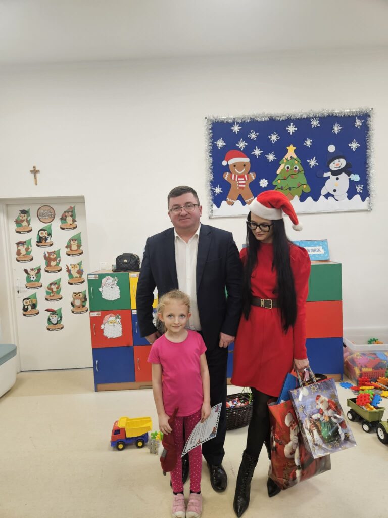 Na zdjęciu są trzy osoby w przedszkolu, na tle kolorowych szafek i zimowej dekoracji. Dziewczynka w różu, kobieta w czerwonej sukience i czapce Mikołaja trzyma torbę z prezentami. Mężczyzna w garniturze stoi za nimi na