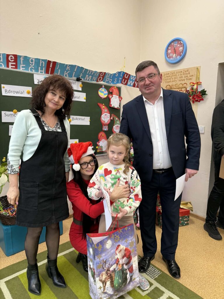 Na zdjęciu są trzy dorosłe osoby i jedno dziecko z dużą torbą z Mikołajem. Tło to szkolna tablica z dekoracjami świątecznymi. Sala jest w jasnych kolorach, z zegarem na ścianie.