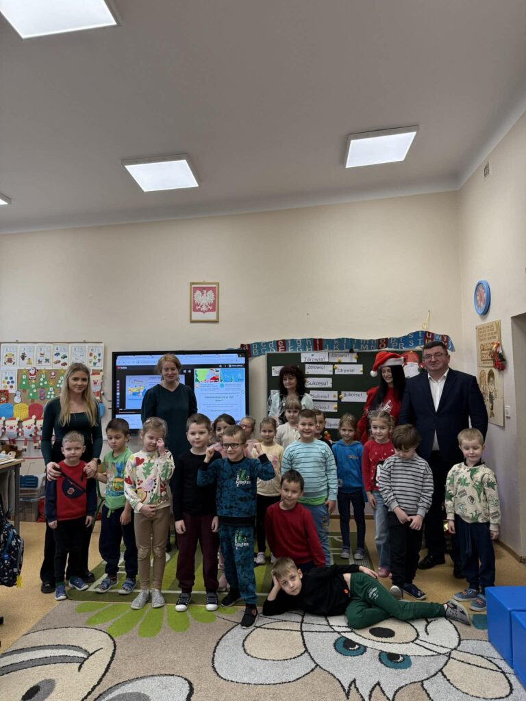 Grupa dzieci i dorosłych w klasie, na dywanie z rysunkiem lwa. Jasne ściany z rysunkami w tle, tablica z kolorowymi literami.