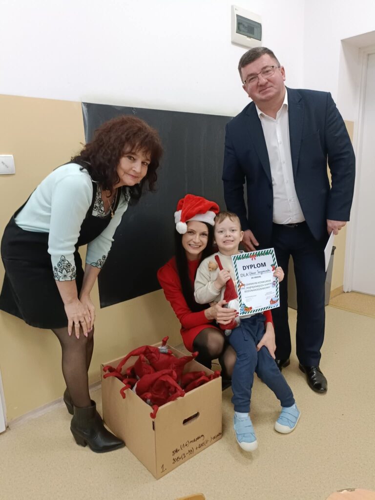 Czworo ludzi w pokoju, kobieta w czapce mikołajowej trzyma dziecko z dyplomem. Obok karton z czerwonymi czapkami. Tło jasne.