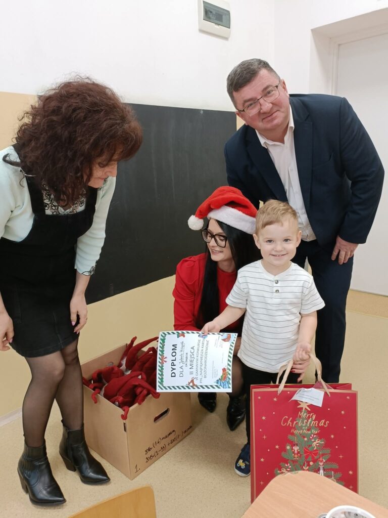 Na zdjęciu jest czworo ludzi, mały chłopiec trzyma dyplom i torebkę świąteczną. W tle są beżowe i czarne ściany. Kobieta w czapce Mikołaja siedzi obok pudła z czerwonymi przedmiotami.