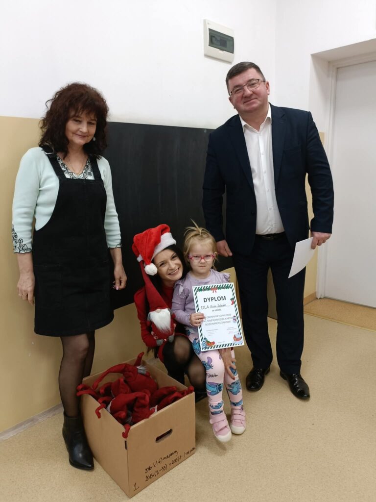 Grupa osób stoi przy ścianie, w tym dziecko z dyplomem. Jedna osoba ma strój Mikołaja, obok znajduje się karton. Tło beżowo-białe z tablicą.