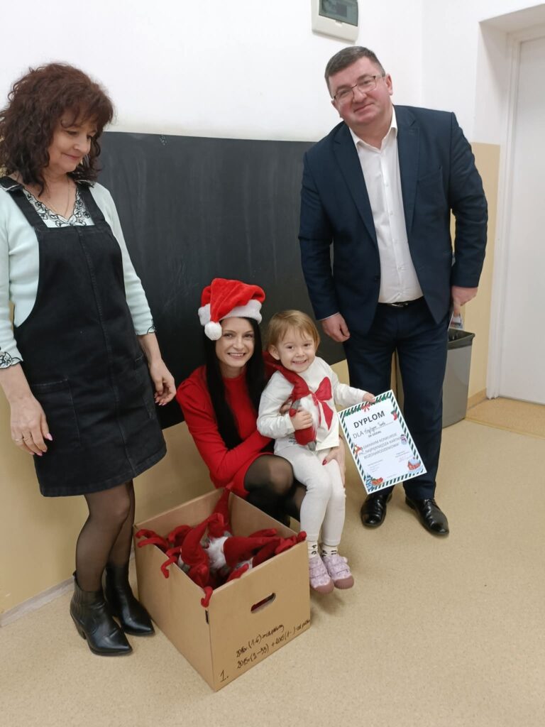 Na zdjęciu widać czworo ludzi, w tym dziecko, z dyplomem w ręce. Kobieta w czapce Mikołaja siedzi obok kartonu z czerwonymi czapkami. Tło jest jasne, z tablicą i beżową ścianą.