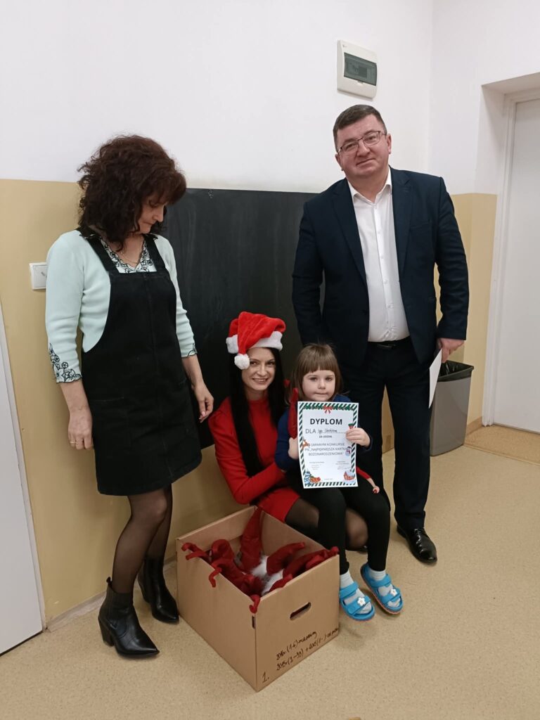 Na zdjęciu widać trzy osoby dorosłe i dziecko, wnętrze z beżowo-białymi ścianami. Dziecko trzyma dyplom, a w tekturowym pudełku leżą czerwone czapki świąteczne. Kobieta w czerwieni ma czapkę M