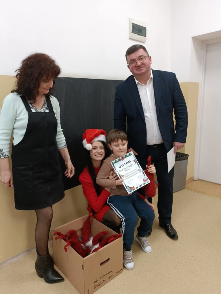 Na zdjęciu widać dwie kobiety, mężczyznę i dziecko w szkole. Kobieta w czerwonym stroju trzyma dziecko z dyplomem. Obok stoi karton z czerwonymi czapkami.