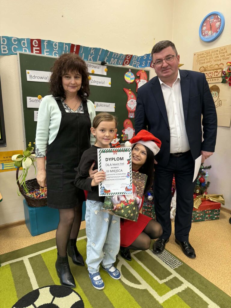 Na zdjęciu są cztery osoby - dziecko trzyma dyplom, otoczone przez dorosłych. Tło stanowi klasa z kolorowymi dekoracjami i zieloną tablicą. Obok znajduje się choinka i kolorowo zapakowane prezenty.