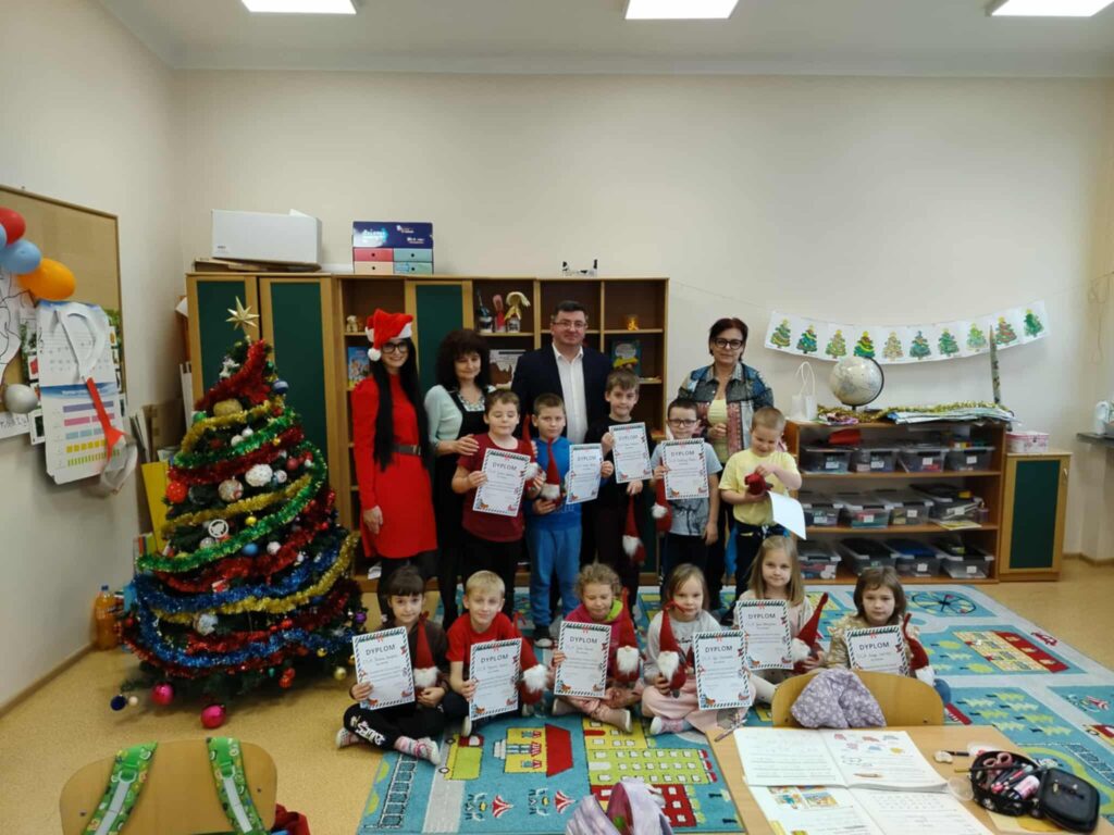 Na zdjęciu grupa dzieci z dorosłymi trzymająca dyplomy, stoi przed ozdobioną choinką w klasie. Tło tworzą szafki i ozdoby świąteczne. Podłoga pokryta jest kolorowym dywanem, a na biurkach leżą przybory