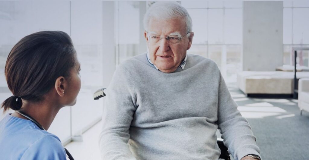 Na zdjęciu jest starszy mężczyzna i młodsza kobieta w niebieskim uniformie medycznym. Rozmawiają w jasnym pomieszczeniu z dużymi oknami. Mężczyzna siedzi na wózku inwalidzkim, ubrany w szary sweter.