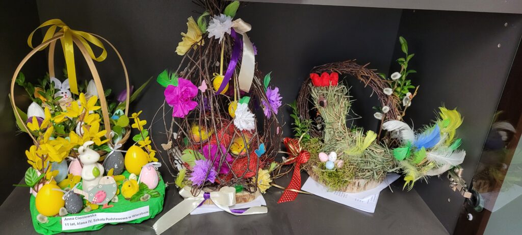 Trzy wielkanocne dekoracje z gałązek, kwiatów i kolorowych pisanek na ciemnym tle. Dominują żółte, zielone i fioletowe akcenty. Zajączki, kurczaczki i piórka dodają świątecznego klimatu.