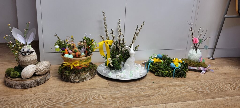 Na zdjęciu widać wielkanocne dekoracje na drewnianej podłodze. Są to koszyki z pisankami, bazie i figurki zajączków. Kolory: zielony, żółty, różowy, biały.