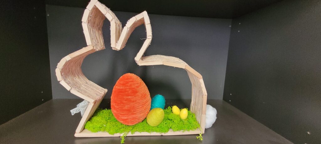 Dekoracja w kształcie zająca z jasnego drewna. We wnętrzu kolorowe jajka na zielonym mchu. Tło ciemne, stonowane.