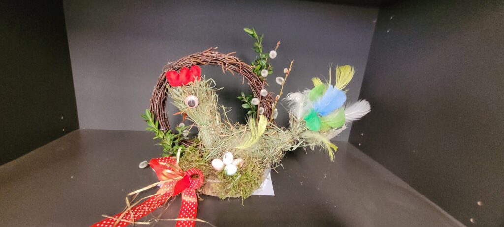 Ręcznie wykonany stroik wielkanocny z wikliny, zielonego mchu i kolorowych piór. Ozdobiony czerwonymi i żółtymi wstążkami. Na tle ciemnoszarej ściany.