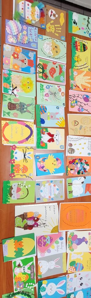 Kolorowe kartki wielkanocne leżą na drewnianym stole. Przedstawiają zajączki, pisanki i kurczaczki. Tło w odcieniach zieleni, żółci i pomarańczy.