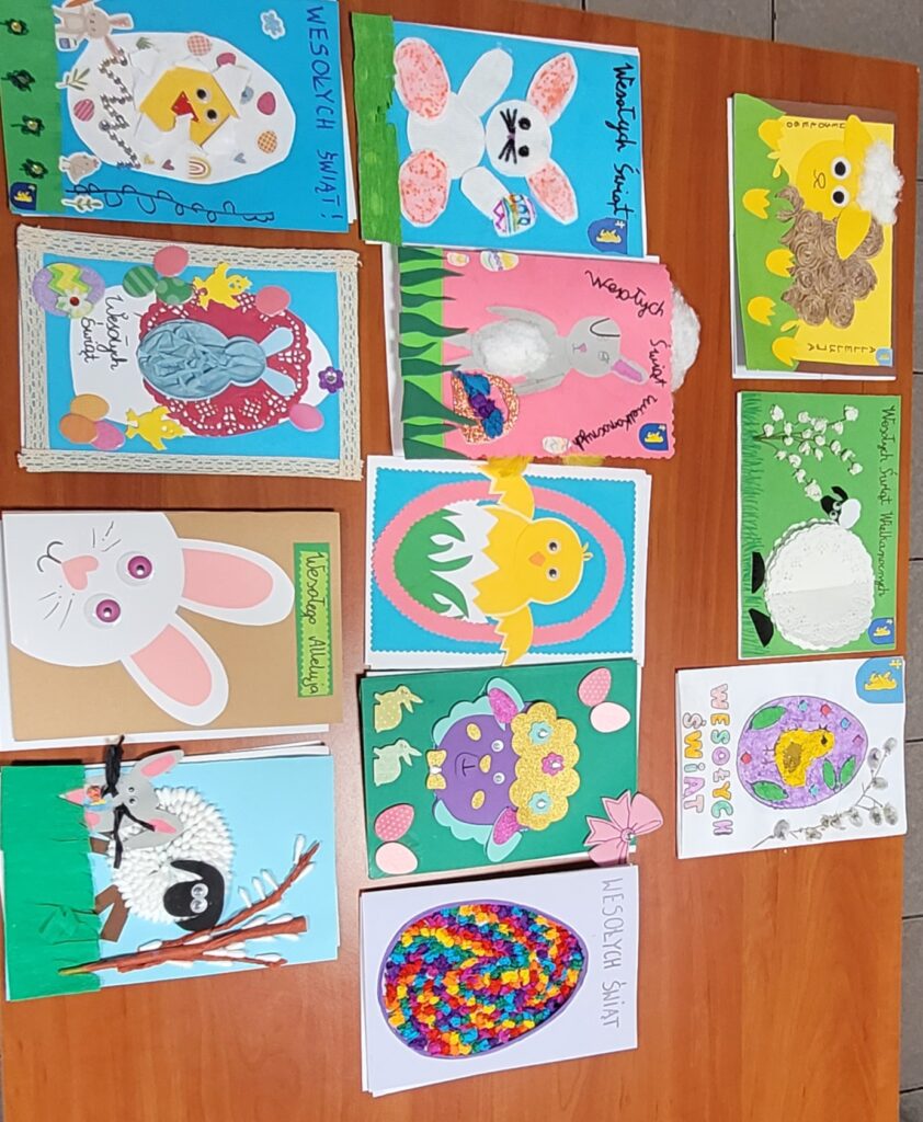 Na zdjęciu widać kolorowe kartki świąteczne z motywami wielkanocnymi, takimi jak króliczki i kurczaczki. Tło stanowi drewniany blat, na którym leżą kartki w różnych kolorach, w tym niebieskim, różowym i zielonym. Kartki oz