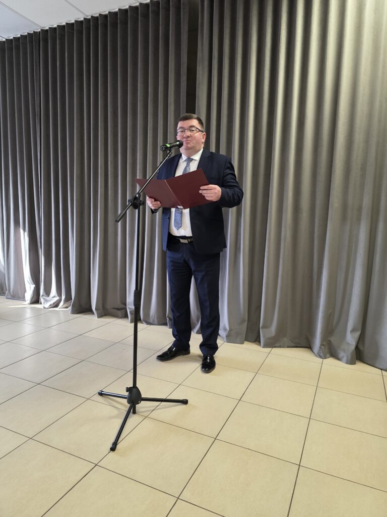 Mężczyzna w garniturze przemawia przy mikrofonie. Tło stanowią szare zasłony. Podłoga jest jasna, kafelkowa.