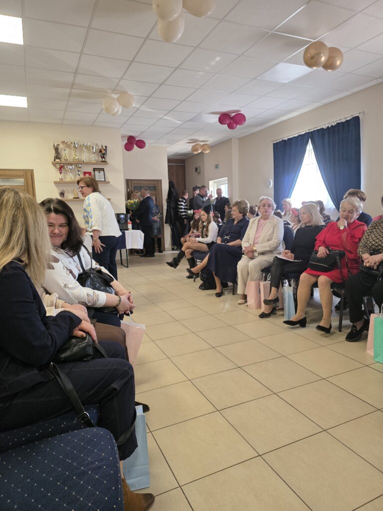 Pomieszczenie z siedzącymi ludźmi na krzesłach, pastelowe ozdoby na suficie. Jasne, kafelkowe podłogi, niebieskie zasłony po prawej. W tle stojący goście i półka z trofeami.