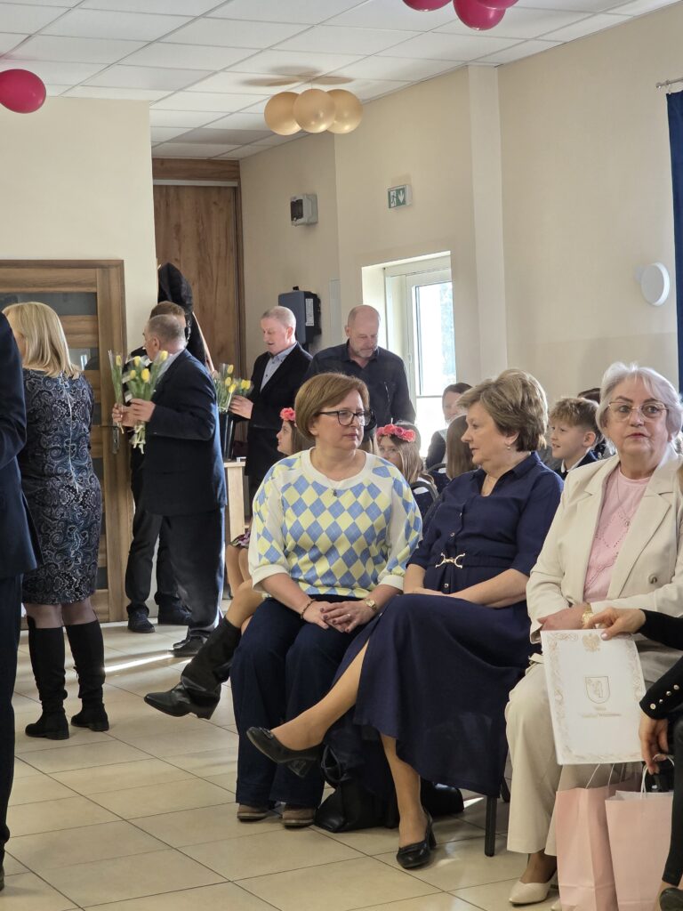 Grupa osób siedzi w jasnym pokoju z balonami w kolorze różowym i złotym. Mężczyźni stoją z żółtymi kwiatami z tyłu. Na ścianie widać drewniane drzwi i okno.