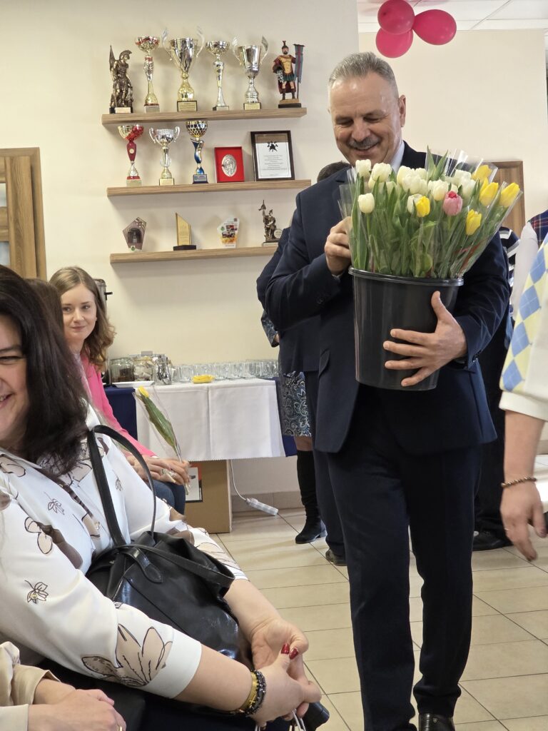 Mężczyzna w garniturze trzyma bukiet tulipanów w jasnych kolorach. W tle półki z trofeami i różowymi balonami. Uśmiechnięci ludzie siedzą przy stole.